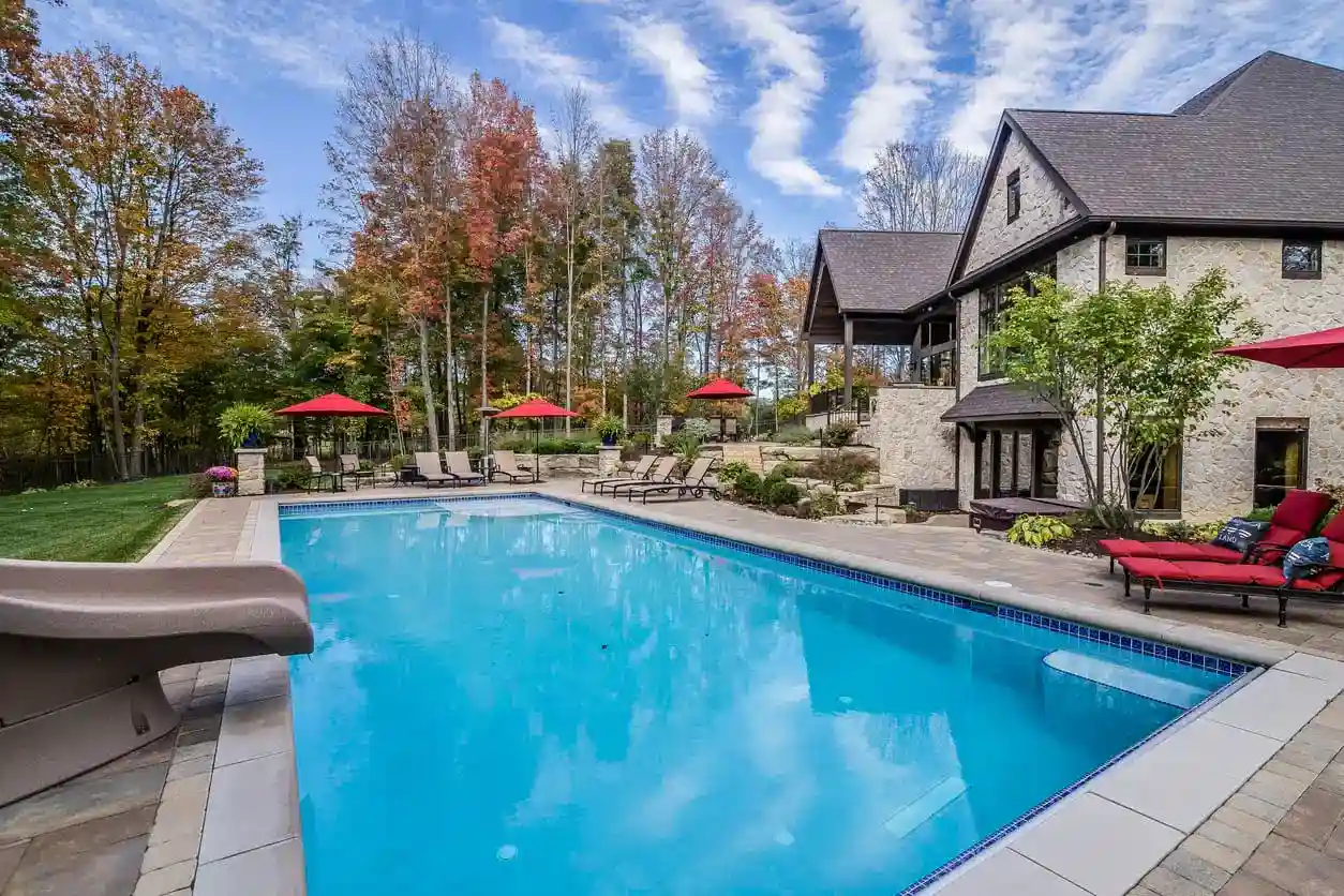 Large rectangular outdoor swimming pool by a stone house, with lounge chairs and red umbrellas—courtesy of Patriot Pool and Spa. Autumn trees frame the scene beneath a partly cloudy sky.