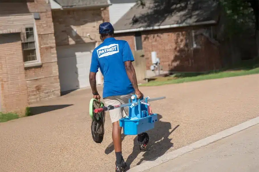 A person wearing a blue "Patriot Pool and Spa" shirt and cap walks on a driveway carrying a blue plastic caddy and other cleaning supplies near a brick house on a sunny day.